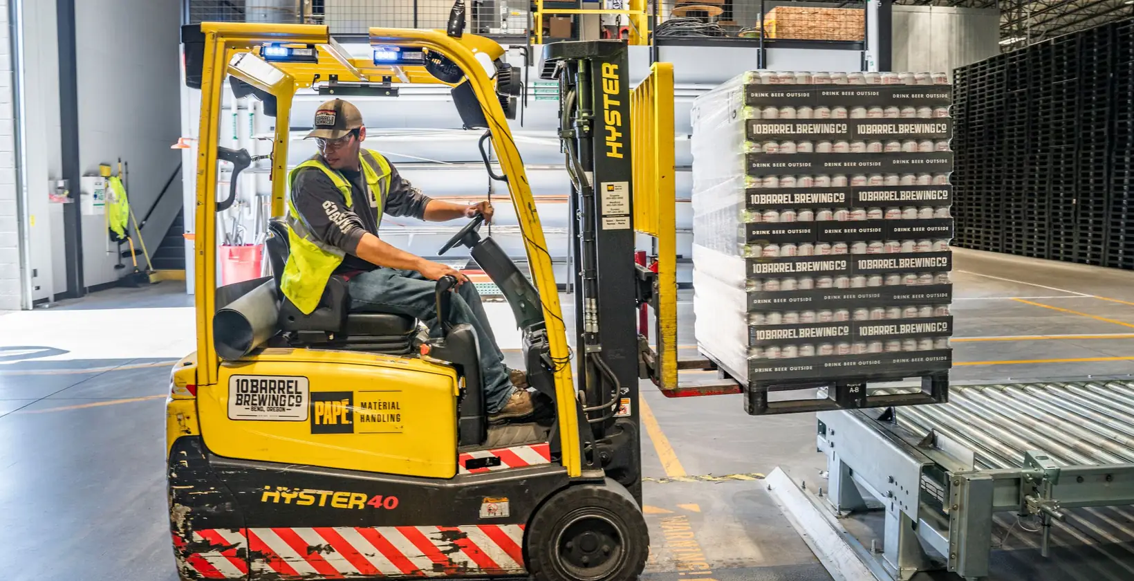 Forklift operator in a hi-vis vest moving a pallet of cases onto a conveyor in a warehouse