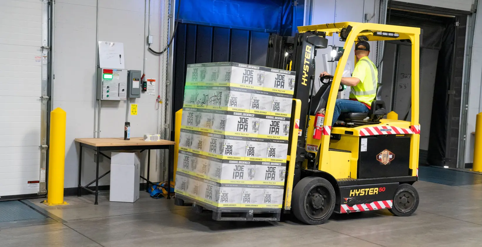 Operator in a hi-vis vest driving a yellow electric forklift loaded with cases at a warehouse dock door